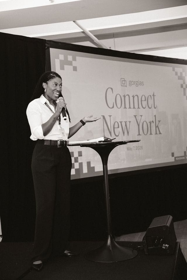 A speaker holding a microphone at a cocktail table by the Connect New York screen, in black and white