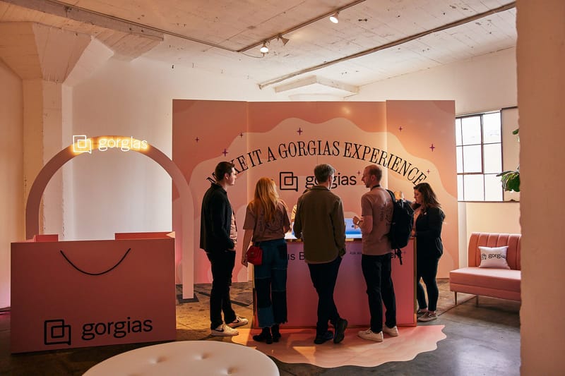 Pink display reading Make It a Gorgias Experience, with a neon Gorgias arch and shopping bag counter