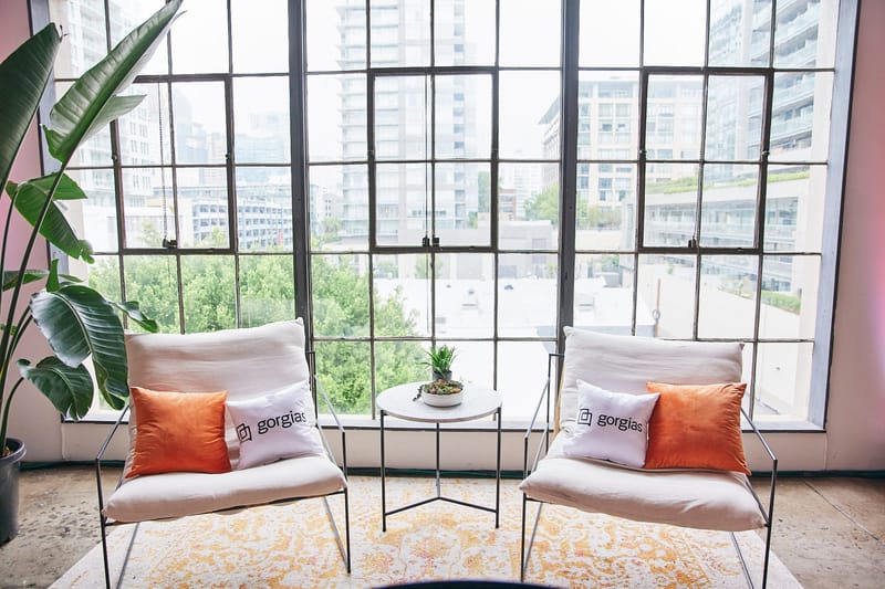 Two white lounge chairs with orange and Gorgias pillows beside a window