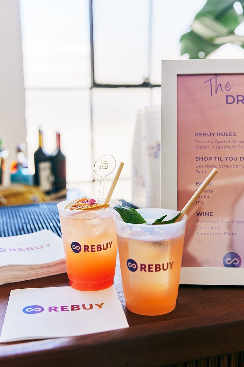 Two Rebuy cocktails beside a drink menu listing Rebuy Rules and Shop Til You Drop