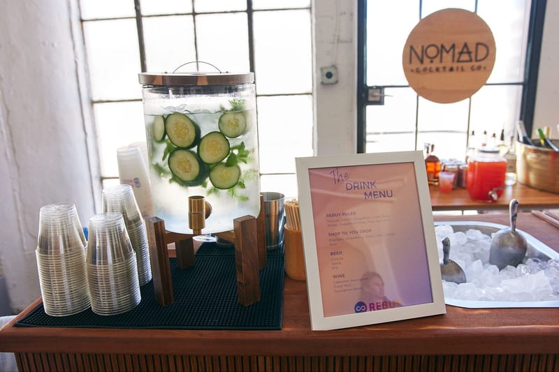 Cucumber water dispenser and cups beside a drink menu frame and a NOMAD Cocktail Co. sign