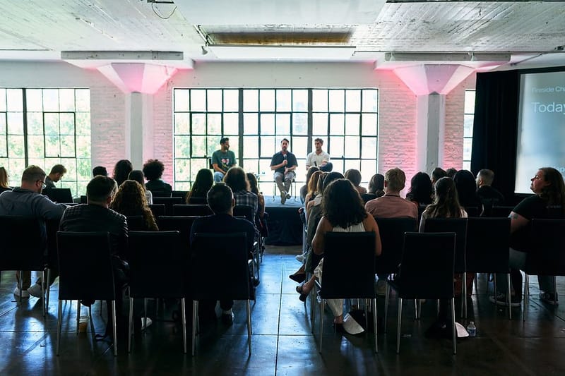 Audience in rows watching three seated panelists in front of steel-framed windows and pink-lit columns