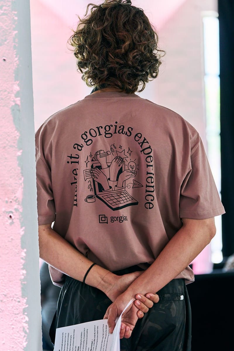 Back of a mauve T-shirt printed make it a gorgias experience beside a pink-lit column