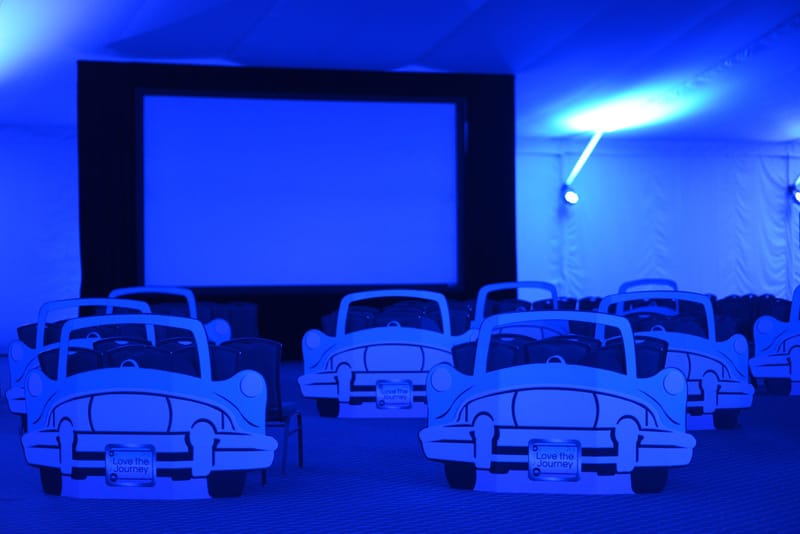 White car cutouts with Love the Journey plates lined up before a large screen in a tent lit blue
