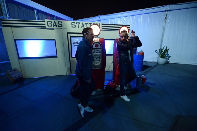 Two people in dark jackets posing beside the red gas pumps at the gas station corner