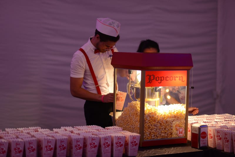 Person in a white paper hat, red suspenders and bow tie scooping popcorn at a red Popcorn machine