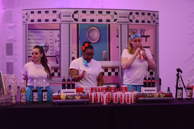 Three people behind a counter of bottles and cups before a diner backdrop signed Milk Shakes, Cheeseburgers and Soda Pop