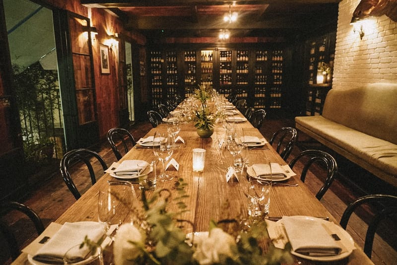 Long wooden dinner table with bentwood chairs, greenery and candles in a room lined with wine racks