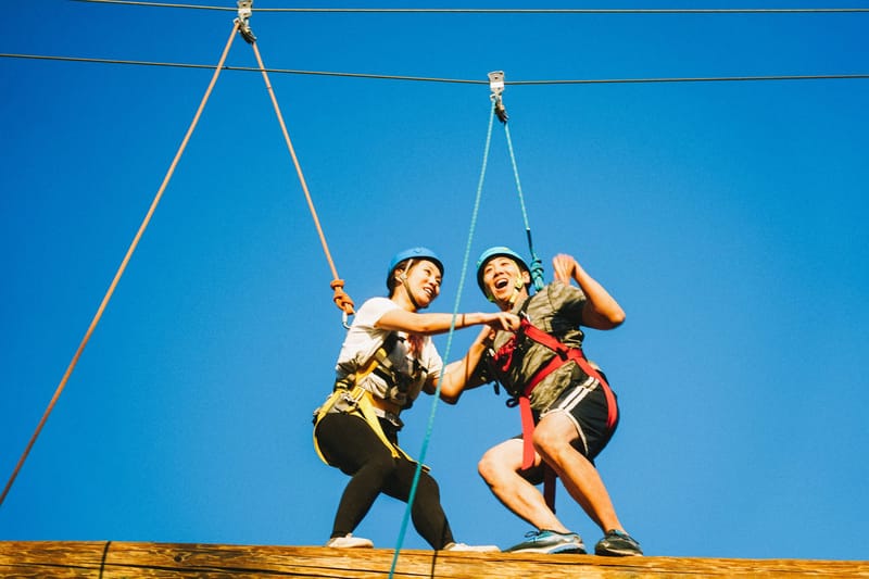 Two guests in helmets and harnesses balancing together on a high wooden beam, clipped to overhead cables