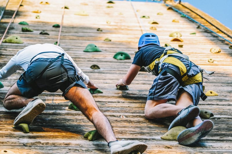 Two guests in harnesses climbing a wooden wall studded with green and yellow holds
