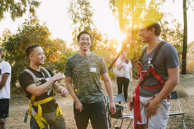 Three smiling guests with name tags, two of them in harnesses, talking outdoors in low evening sun