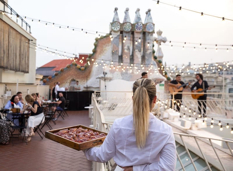 Server carrying a tray of bites across the roof terrace toward guests and guitarists under string lights