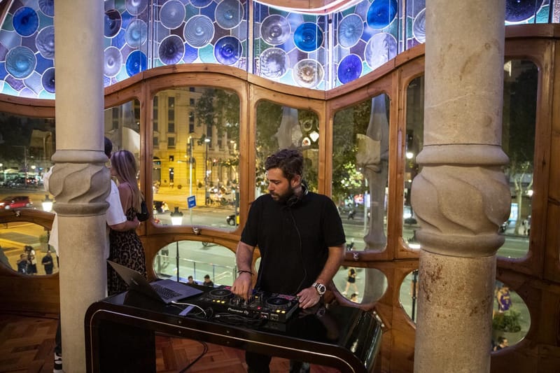 A DJ at decks in front of curved wooden windows with blue glass roundels and a street view