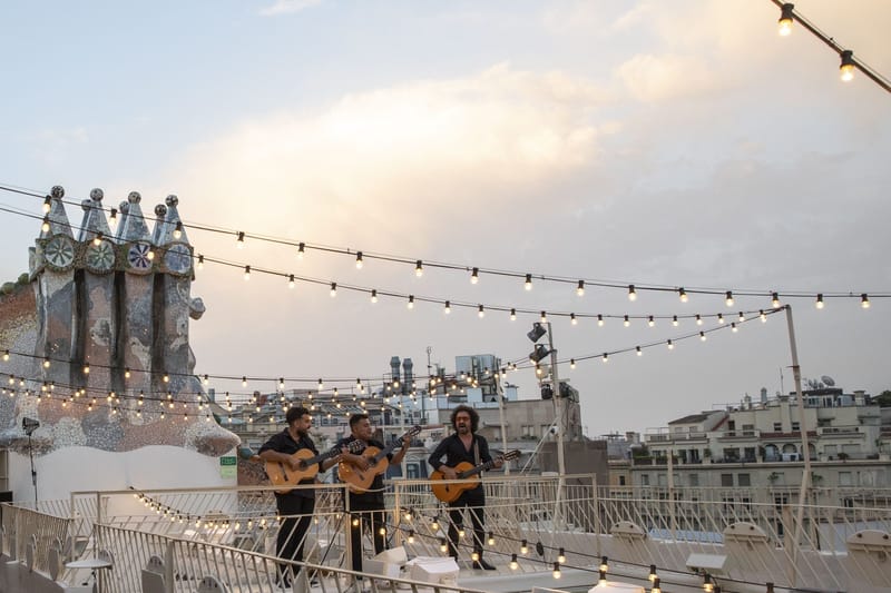 Three guitarists playing on the rooftop at dusk under strings of lights