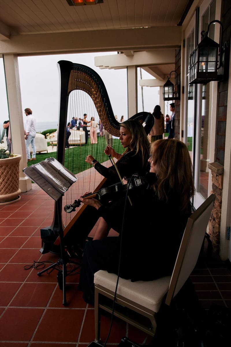 Harpist and violinist playing on a tiled covered terrace while guests gather on the lawn outside