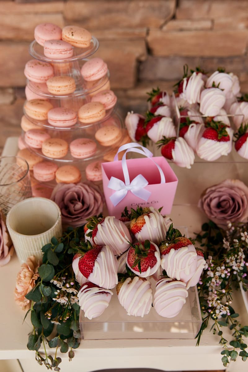 Macaron tower beside pink and white dipped strawberries, a pink gift bag and dusty rose blooms