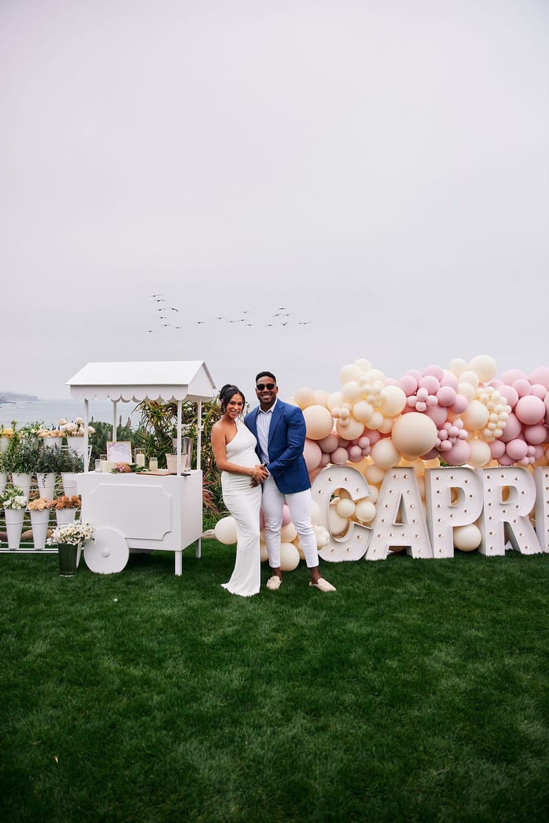 Expecting mother and a man in a blue blazer on the lawn by a white flower cart and CAPRI letters