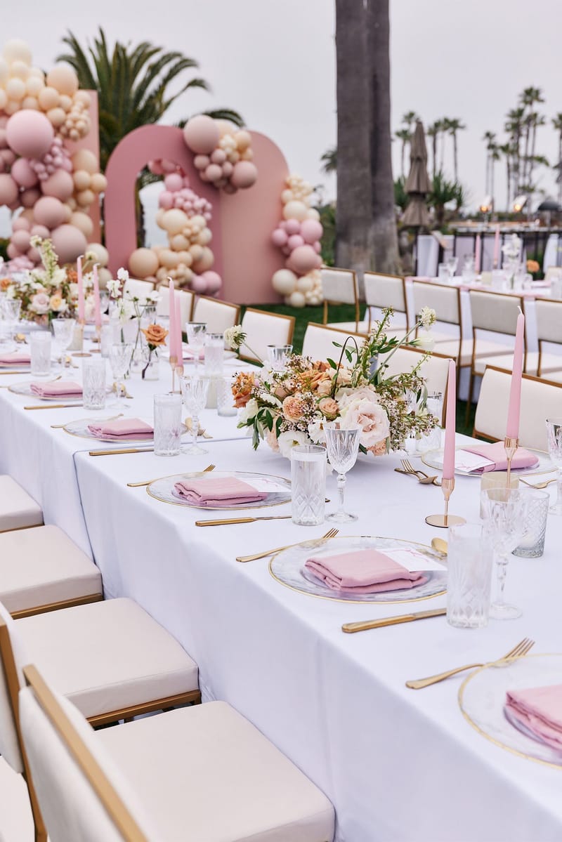 Long white table with pink napkins, pink taper candles and peach florals, pink arched balloon backdrops at the end