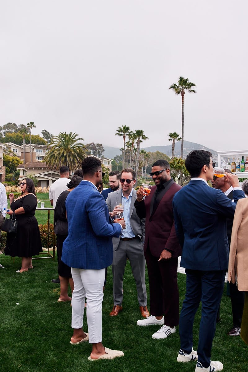 Guests in blazers and dresses mingling on a lawn with palm trees behind