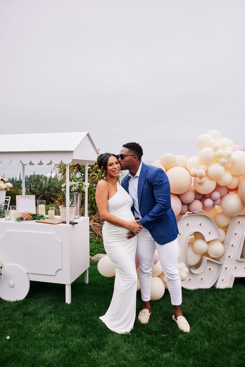 Expecting mother and a man in a blue blazer kissing beside a white flower cart and CAPRI letters