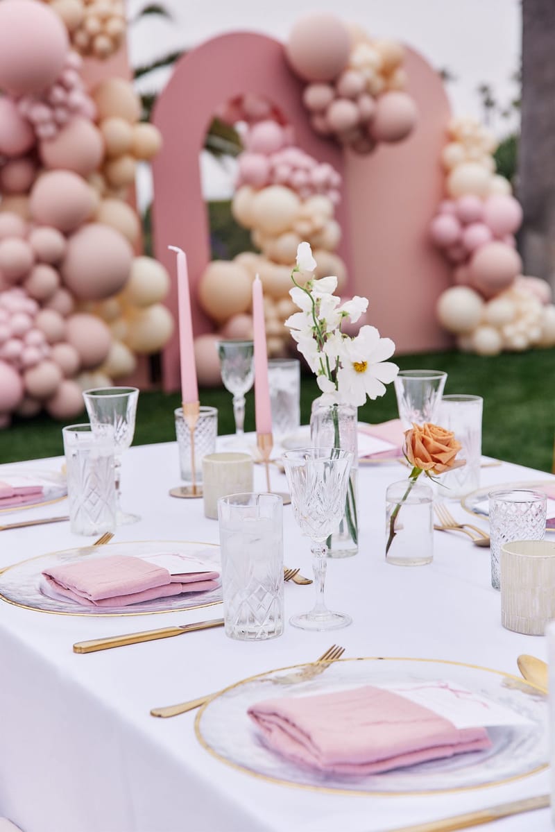 Place settings with pink napkins, gold flatware and pink tapers in front of a pink arched balloon backdrop