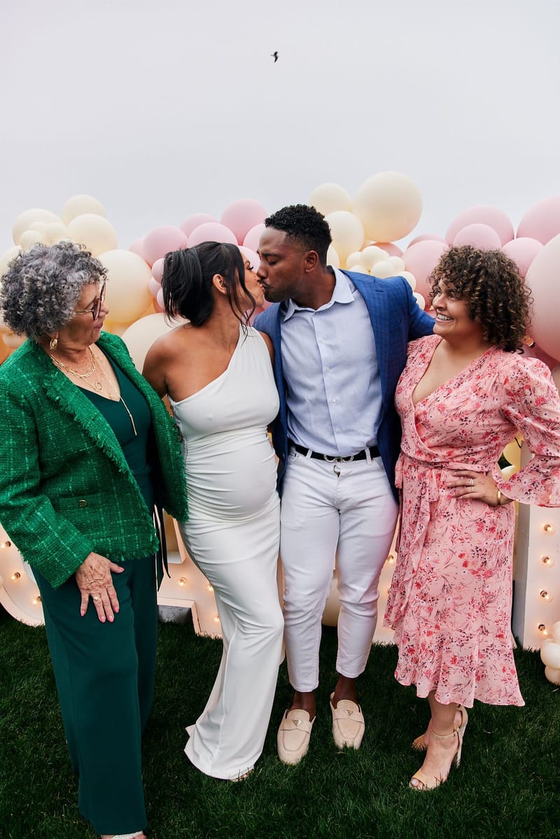 Expecting mother and a man in a blue blazer kissing between two women in green and floral dresses
