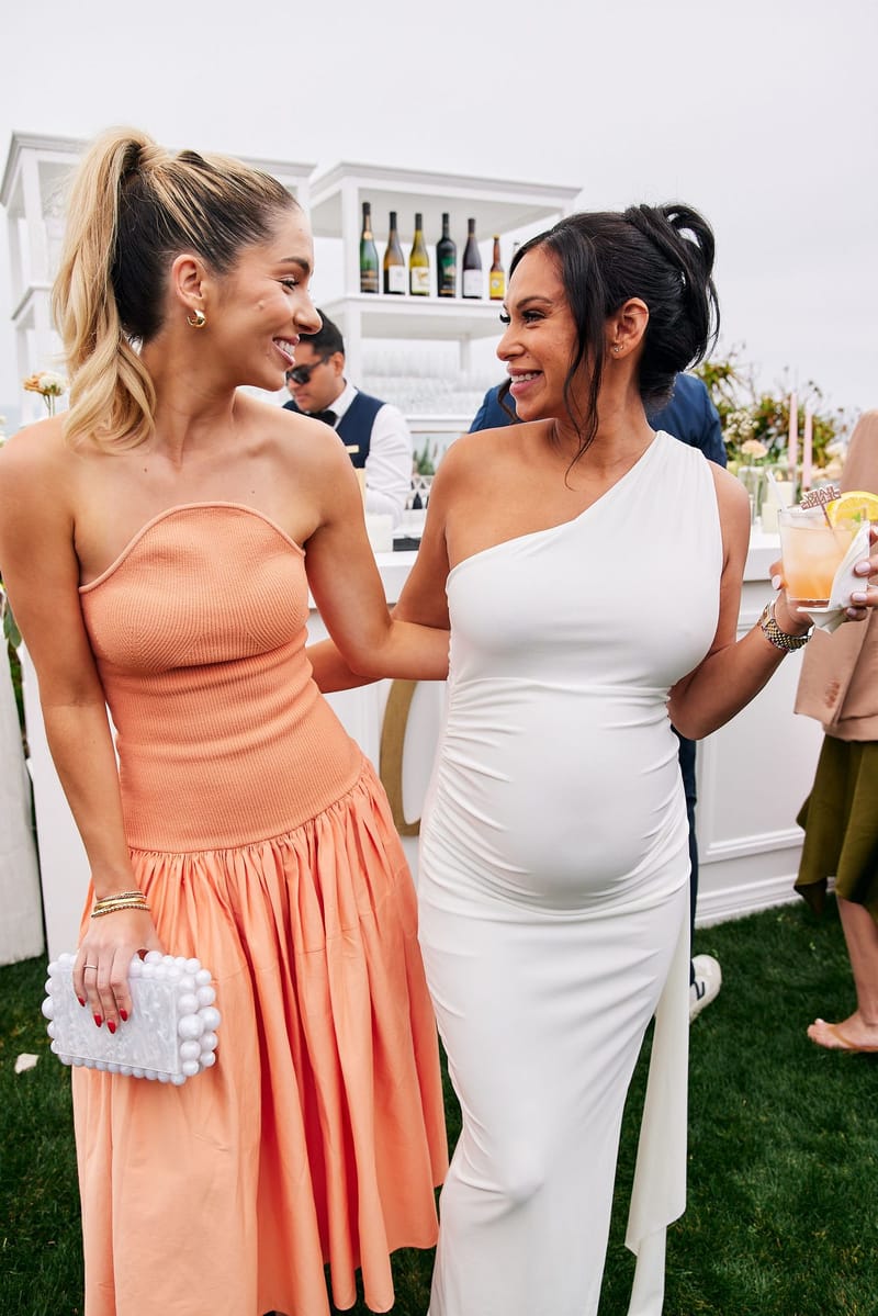Expecting mother in a white one-shoulder dress laughing with a guest in an orange dress