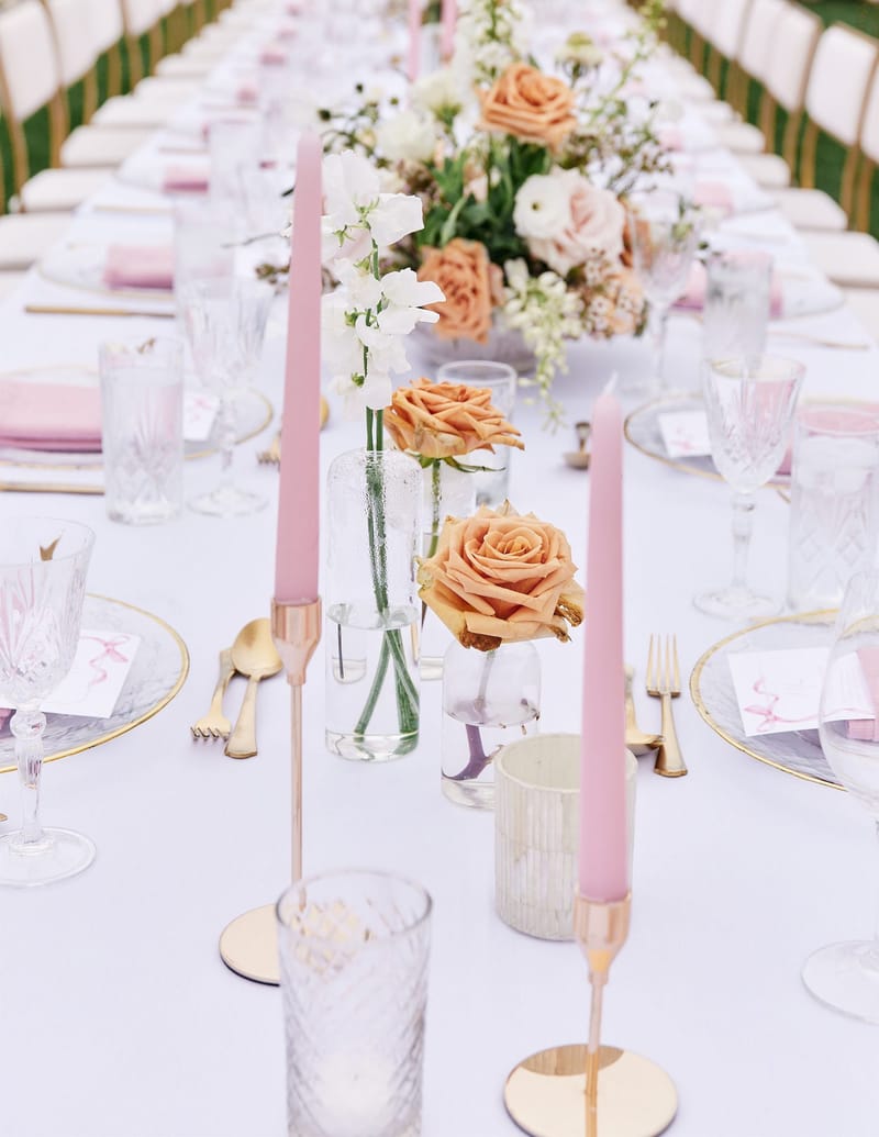 Close view down a long table with pink taper candles, peach roses, crystal glasses and gold flatware