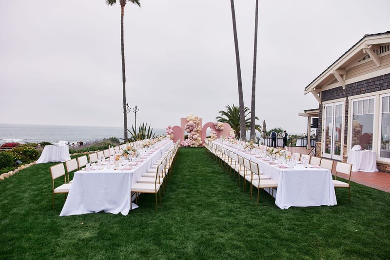Two long white banquet tables on a lawn by the ocean and palms, leading to pink arched balloon backdrops