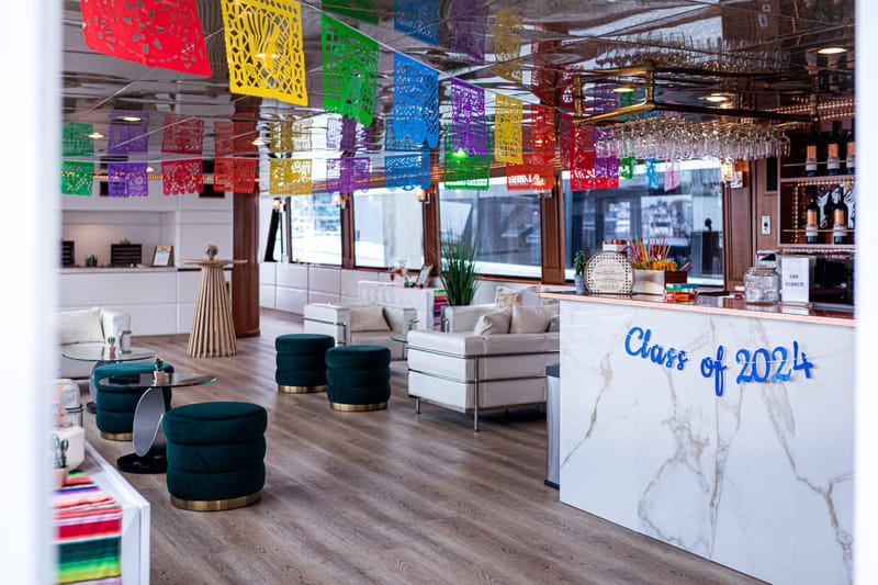 Lounge under colorful papel picado with teal velvet ottomans, white sofas and a marble bar reading Class of 2024
