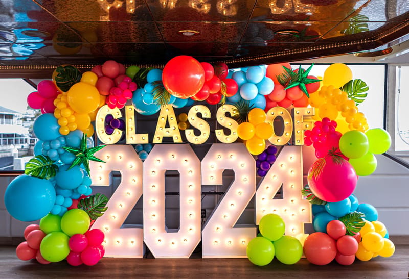 Light-up CLASS OF 2024 marquee letters set in a colorful balloon garland with tropical leaves