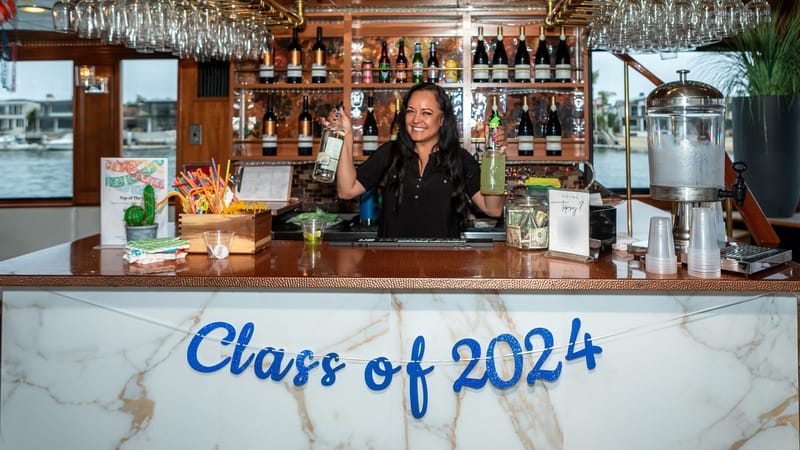 Smiling bartender holding up two bottles behind a marble bar with a blue glitter Class of 2024 banner