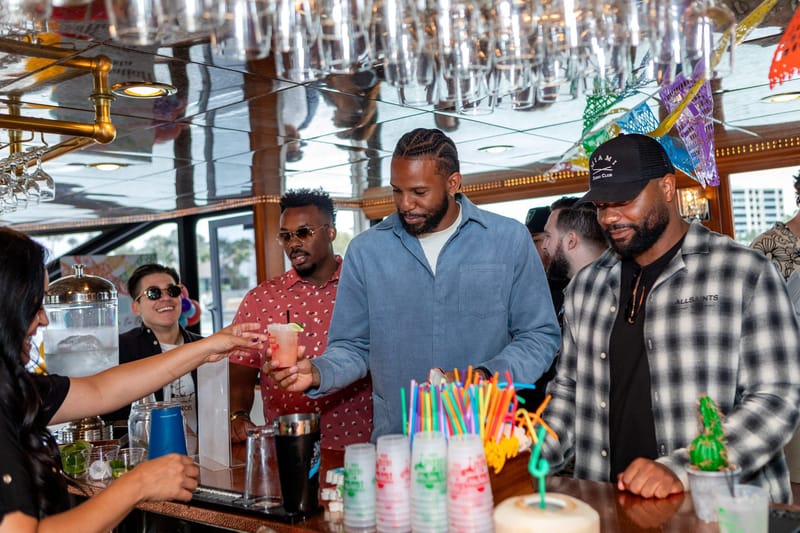 Bartender handing a pink drink with lime to a guest at the bar, with colorful straws and stacked cups