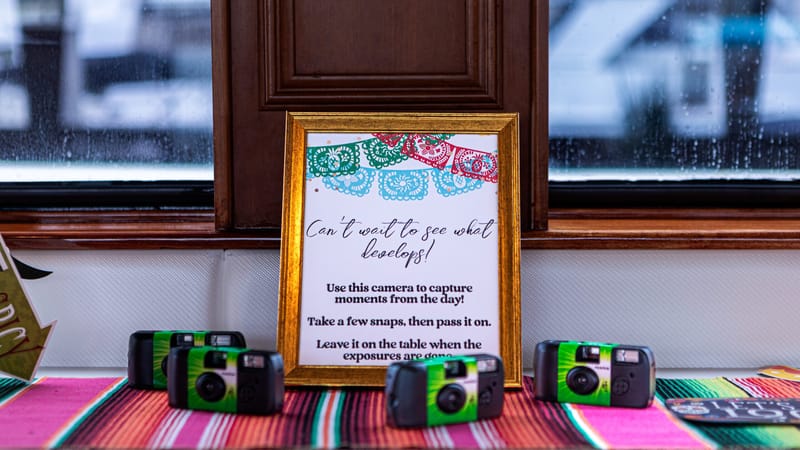 Gold-framed sign reading Can't wait to see what develops! behind green disposable cameras on a serape runner
