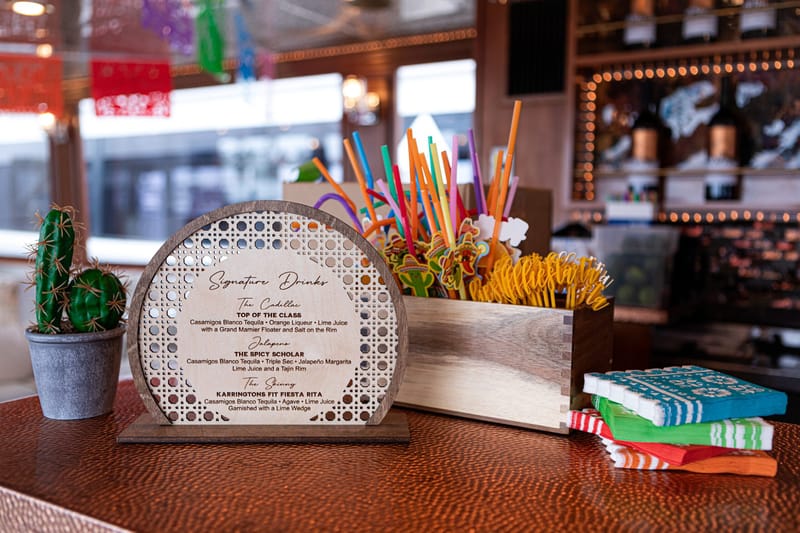 Round wooden Signature Drinks menu listing Top of the Class and The Spicy Scholar beside a crate of colorful straws