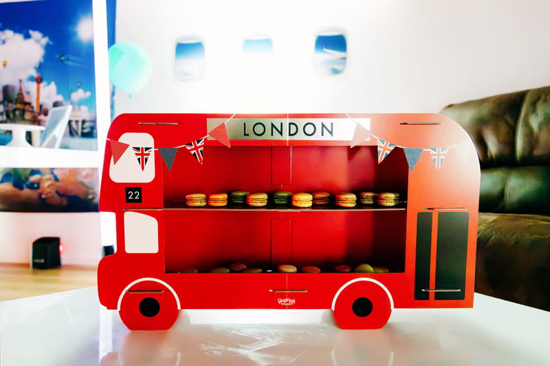 Red London double-decker bus stand holding a row of macarons, with airplane window decals on the wall