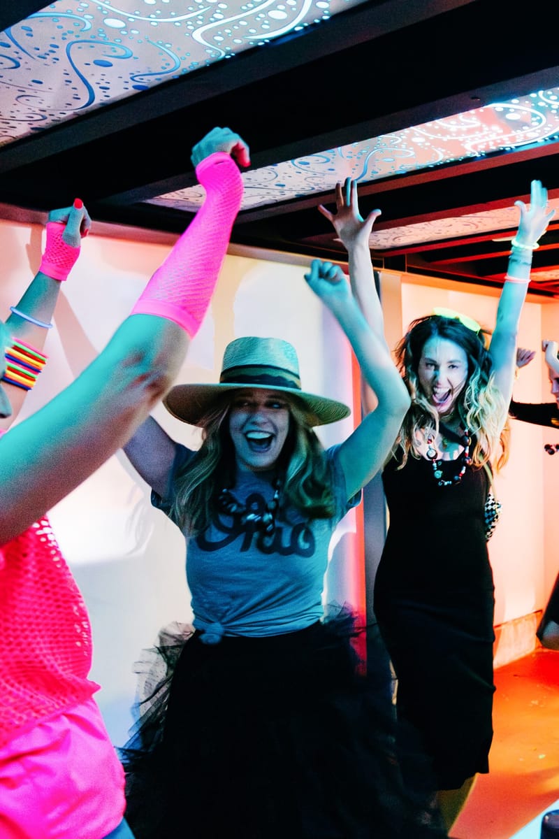 Two women dancing with arms raised in neon 80s outfits, one wearing a wide-brim hat