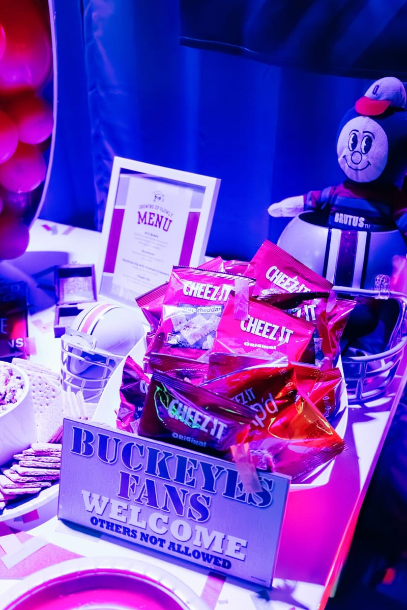 Basket of Cheez-It packs behind a Buckeyes Fans Welcome sign, with football helmets under purple light