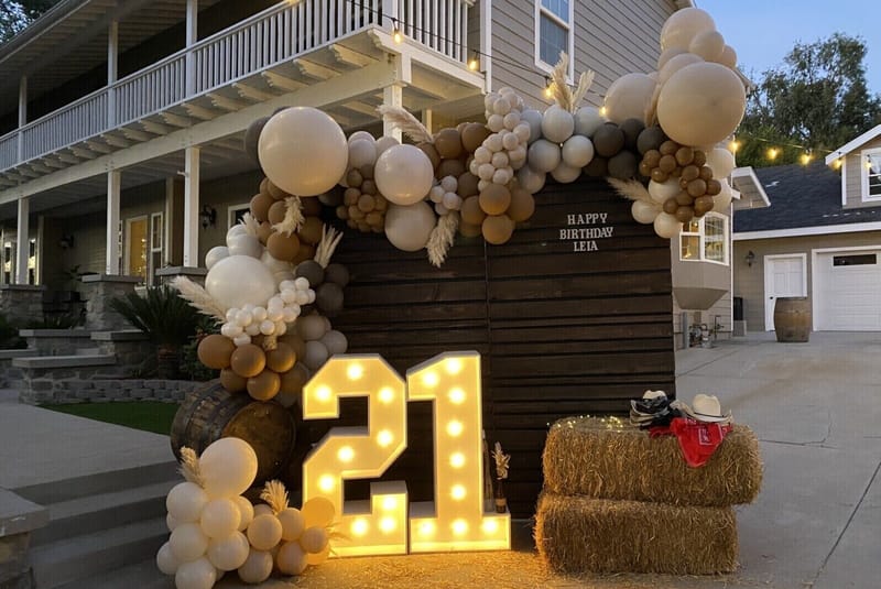 Lit 21 marquee numbers, a Happy Birthday Leia plank wall with a balloon garland, and hay bales in a driveway