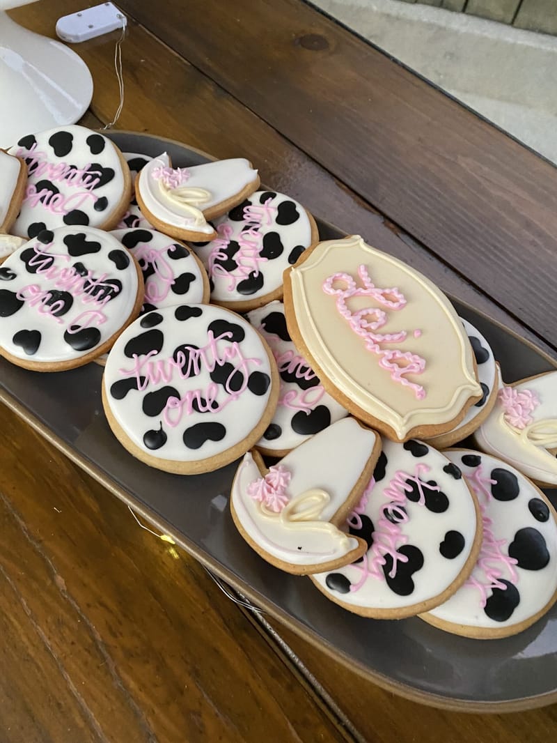 Tray of cow print cookies piped with twenty one in pink and a cream plaque cookie piped with Leia