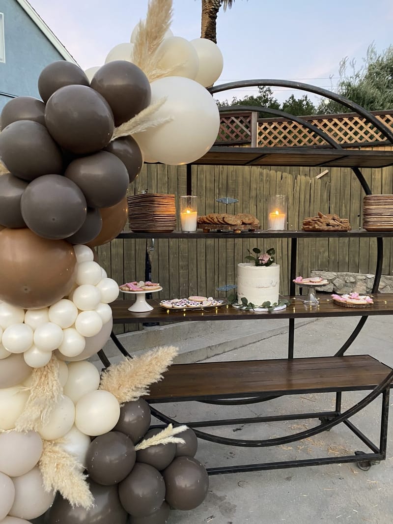 Dessert shelf with a small white cake, cookies and candles, framed by brown and cream balloons with pampas grass