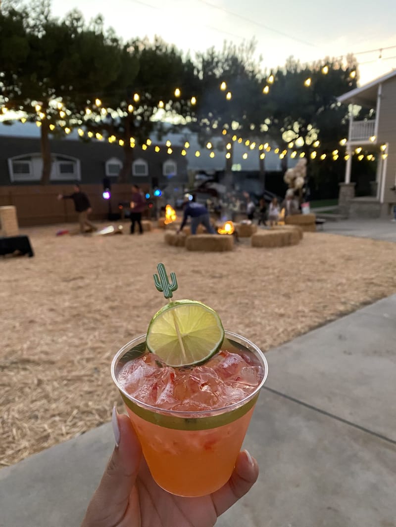 A hand holding a drink with a lime wheel and cactus pick over a string-lit yard of hay bales