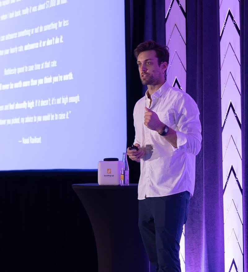 Speaker in a white shirt presenting beside a quote slide and a Leveling Up cube on a cocktail table