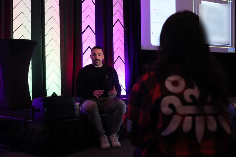 Speaker seated in front of lit chevron pillars as a guest watches from the foreground