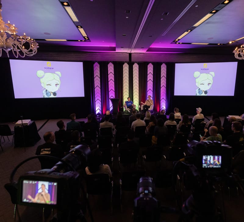 Two speakers in armchairs under screens showing the Leveling Up logo and a cartoon character