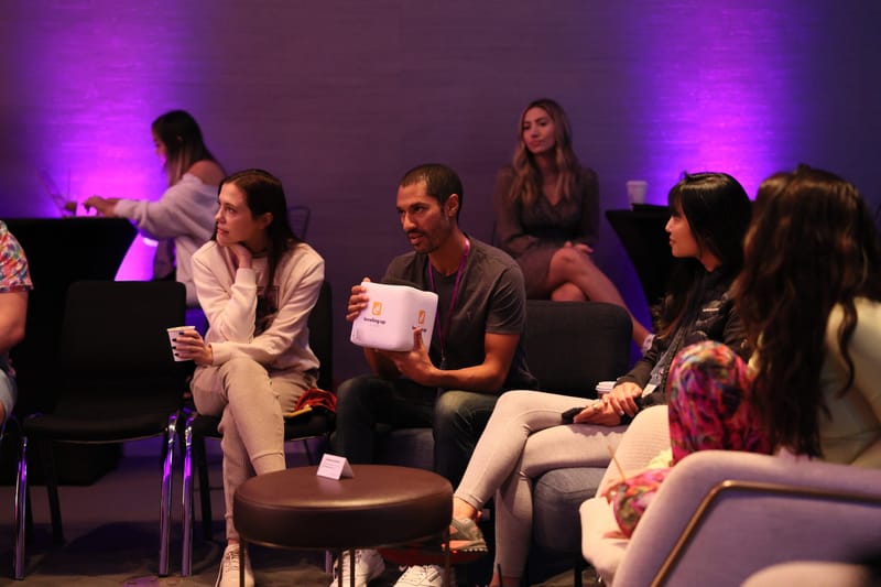 Guests in lounge chairs around small round tables, one holding a cube microphone