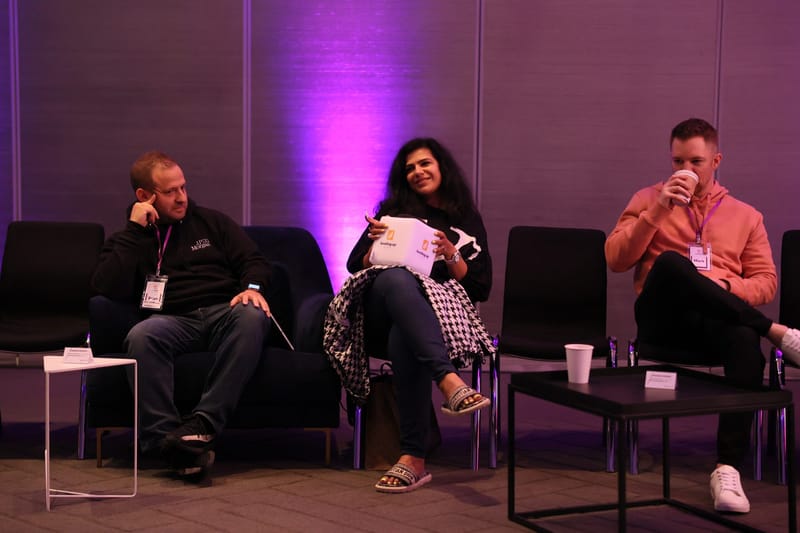 Three guests with lanyards in lounge chairs against a purple-lit wall, one holding a cube microphone