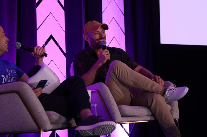 Two speakers laughing in armchairs with microphones in front of purple chevron panels