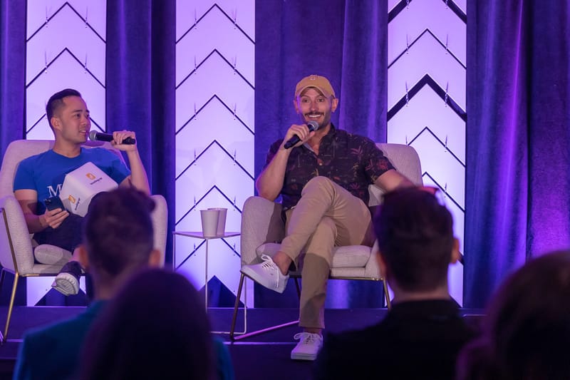 Two speakers with microphones in cream armchairs on the purple-lit stage
