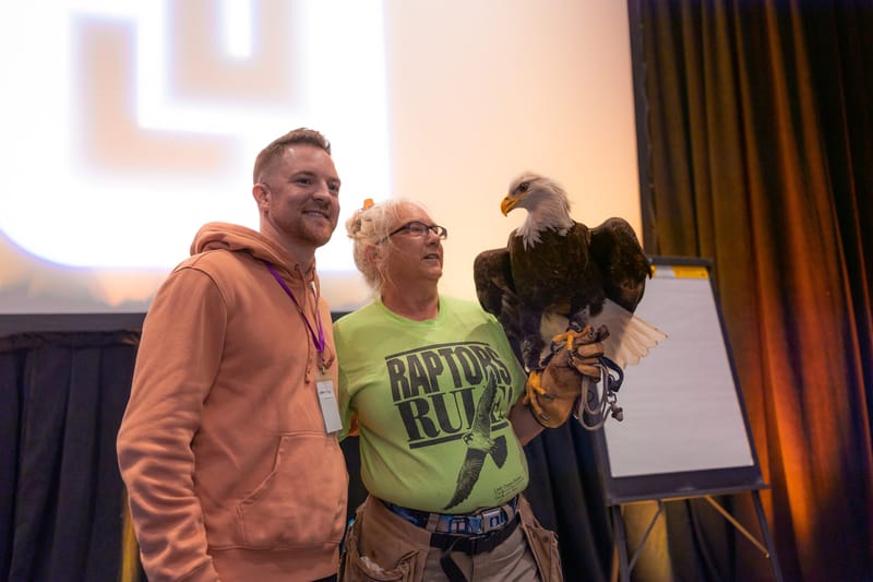 A bald eagle perched on a handler's glove beside a smiling guest in a pink hoodie